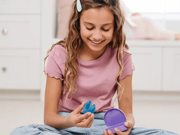 A smiling girl sitting on her bedroom floor holding her Toothpillow appliance and case, with her Toothpillow kit box