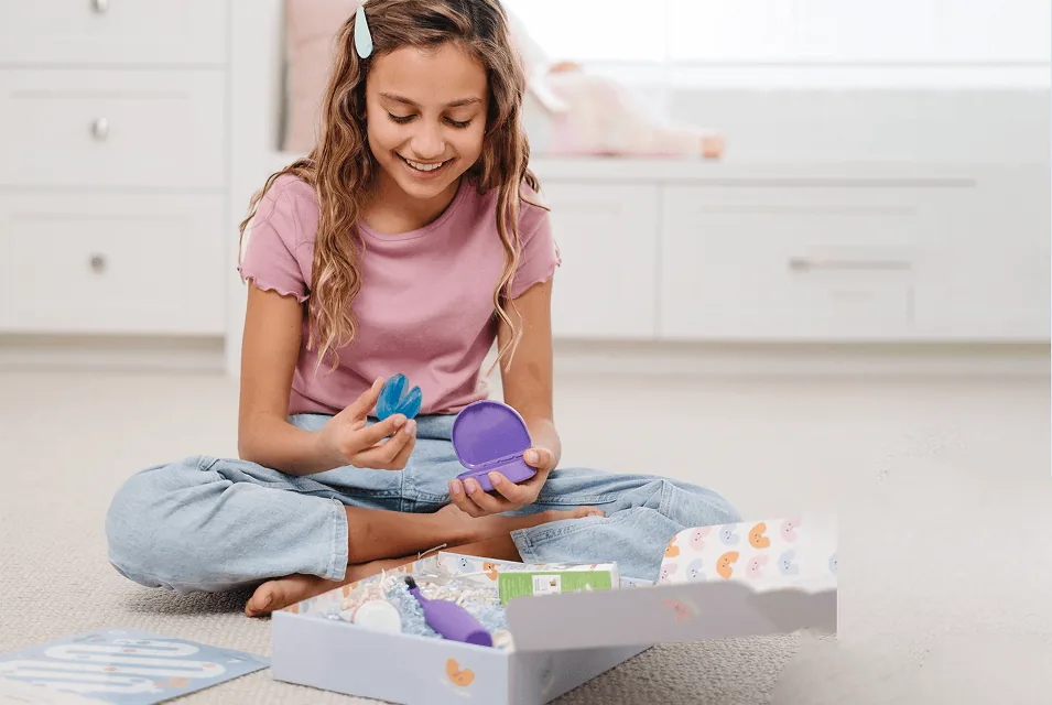 A smiling girl sitting on her bedroom floor holding her Toothpillow appliance and case, with her Toothpillow kit box